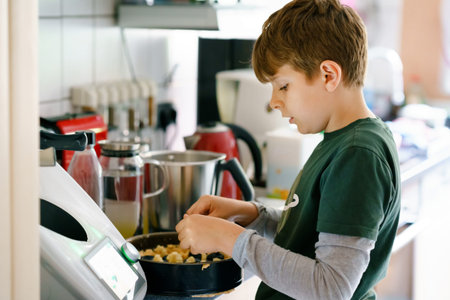Happy blond school kid boy baking plum cake in domestic kitchen, indoors. Funny lovely healthy...