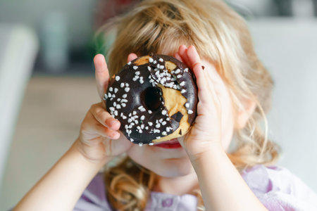 Happy little preschool girl eating sweet donut indoor. Blond child in the city. Cute kid and...