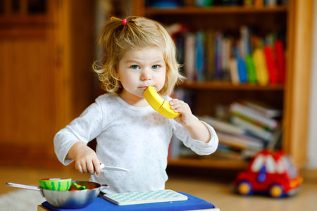 Cute little toddler girl playing at home with eco wooden toys. Happy healthy excited child cutting vegetables and fruits with toy knife. Baby girl having fun with role kitchen and cooking game.の写真素材