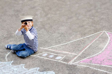Little kid boy as pirate on ship or sailingboat picture painting with colorful chalks on asphalt. Creative leisure for children outdoors in summer. Child with captain hat and binoculars.の写真素材