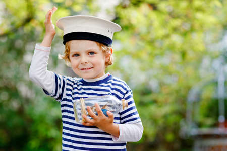 Happy little kid boy in sailor capitain hat and uniform playing with sailor boat ship. Smiling preschool child dreaming and having fun. Education, profession, dream conceptの写真素材