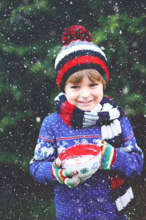 Happy little kid boy drinking hot cocoa and chocolate drink and marshmallows. Funny child in winter sweater, cap, long warm scarf and colorful gloves. On cold winter day during snowfall outdoorsの写真素材