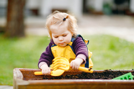 Cute adorable toddler girl playing with sand and shovel on spring day. Baby child wearing yellow boots and mud rain puddle pants. Happy girl planting vegetables in spring.の写真素材