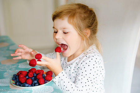 Cute little preschool girl eating fresh raspberries and blueberries. Happy child tasting raspberry and blueberry. Healthy food, childhood and development. Happy kid at home or nursery.の写真素材
