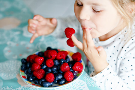 Cute little preschool girl eating fresh raspberries and blueberries. Happy child tasting raspberry and blueberry. Healthy food, childhood and development. Happy kid at home or nursery.の写真素材