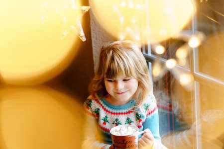 Little preschool girl holding cup with hot chocolate with marhsmallows. Happy child drinking sweet cocoa by window with Christmas lights in winter. Cozy family celebration of xmas.の写真素材