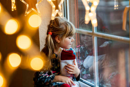 Little preschool girl holding cup Santa Claus boot with gift called Nikolausstiefel in German. Happy child wait on holiday by window with Christmas lights in winter. Cozy family celebration of xmas.の写真素材