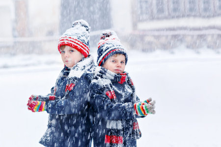 Two happy little kid boys in colorful fashion clothes playing outdoors during strong snowfall....
