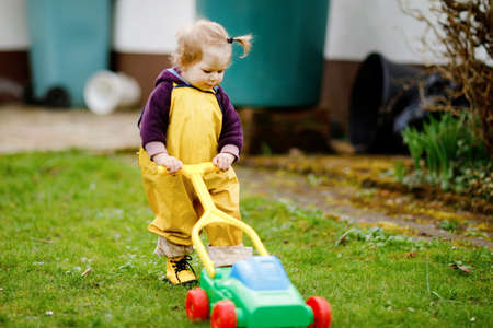 Cute adorable toddler girl playing with toy lawn mower outdoors on spring day. Baby child wearing yellow mud rain puddle pants trousers. Portrait of happy girl.の写真素材