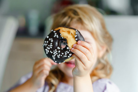 Happy little preschool girl eating sweet donut indoor. Blond child in the city. Cute kid and...