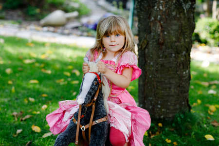 Little preschool girl hugging with rocking horse toy. Happy child in princess dress on sunny summer day in garden. Girl in love with her favourite old vintage toy animal.の写真素材