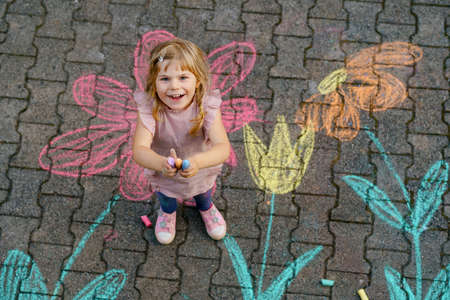 Little preschool girl painting with colorful chalks flowers on ground on backyard. Positive happy toddler child drawing and creating pictures on asphalt. Creative outdoors children activity in summer.の写真素材