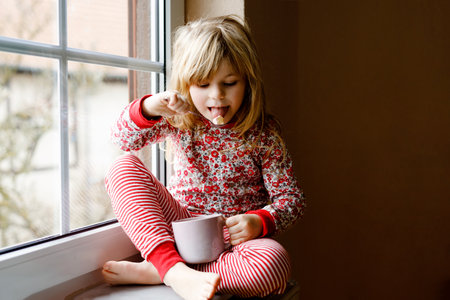 Little preschool girl holding cup with hot chocolate with marhsmallows. Happy child in pajamas drinking sweet cocoa by window in winter. Cozy family celebration of xmas.の写真素材