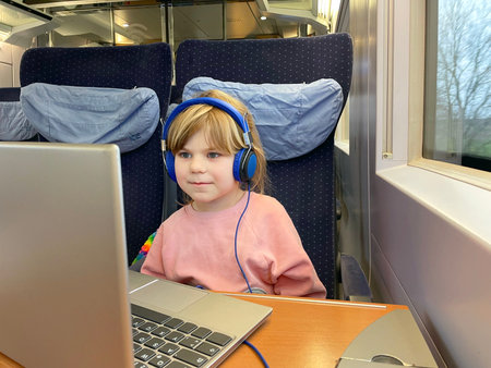 Cute little preschool girl sitting in train and watching tv on laptop while moving. Happy child...