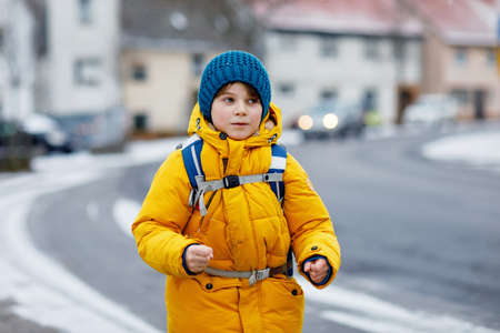 Little school kid boy of elementary class walking to school during snowfall. Happy child having fun and playing with first snow. Student with in yellow jacket and backpack in colorful winter clothes.の写真素材