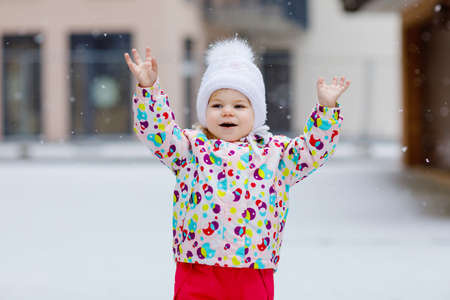 Portrait of little toddler girl walking outdoors in winter. Cute toddler eating sweet lollypop candy. Child having fun on cold snow day. Wearing warm baby colorful clothes and hat with bobbles.の写真素材