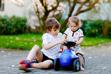 Cute little baby girl and school kid boy playing with blue small toy car in garden of home or nursery. Toddler sister and brother children having fun together.の写真素材