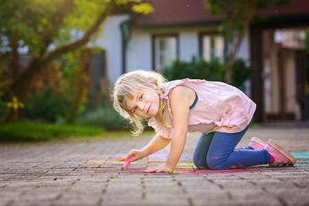 Little preschool girl painting with colorful chalks flowers on ground on backyard. Positive happy toddler child drawing and creating pictures on asphalt. Creative outdoors children activity in summer.の写真素材