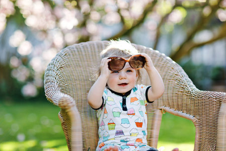 Cute little baby girl sitting on big chair in garden. Beautiful happy smiling toddler with blooming...
