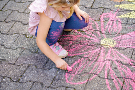 Little preschool girl painting with colorful chalks flowers on ground on backyard. Positive happy...