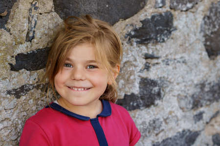 Portrait of happy smiling toddler girl in red clothes outdoors. Little child with blond hairs looking and smiling at the camera. Happy healthy child enjoy outdoor activity and playing.の写真素材