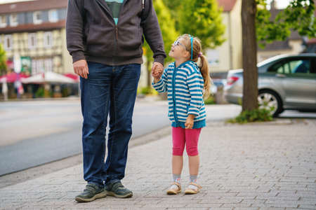 Happy child preschool girl smiles and holds fathers hand on the way to school on the first sunny day of class. Dad and daughter on the way to kindergarten, fun together. Child with glasses.の写真素材