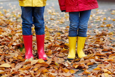 Two little children playing in red and yellow rubber boots in autumn park in colorful rain coats and clothes. Closeup of kids legs in shoes dancing and walking through fall autumnal leaves and foliageの写真素材