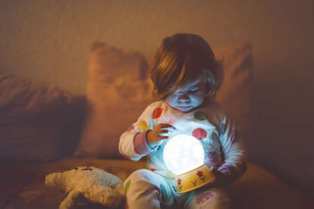 Cute little toddler girl playing with colorful night light lamp before going to bed. Sleepy tired baby daughter in nightwear having fun. Healthy child not sleeping.の写真素材
