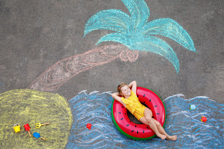 Happy little preschool girl in swimsuit on inflatable ring with sea, sand, palm painted with colorful chalks on asphalt. Cute child with having fun with chalk picture. Summer, vacations, summertimeの写真素材