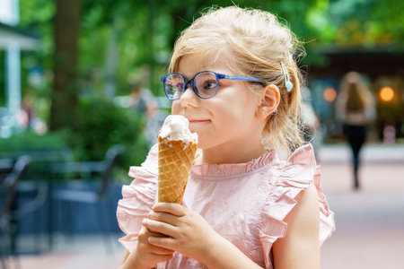 Little preschool girl with glasses eating ice cream in waffle cone on sunny summer day. Happy...