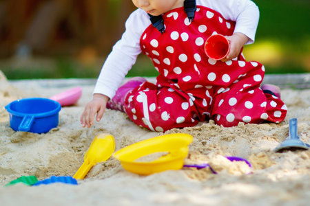 Cute toddler girl playing in sand on outdoor playground. Beautiful baby in red gum trousers having fun on sunny warm summer day. Child with colorful sand toys. Healthy active baby outdoors plays gamesの写真素材