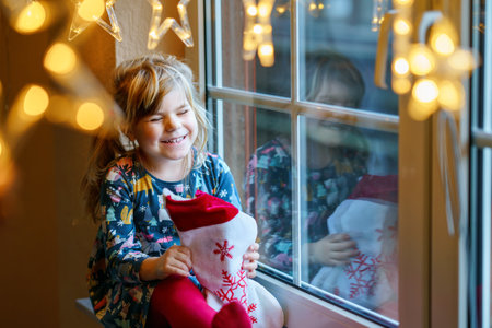 Little preschool girl holding cup Santa Claus boot with gift called Nikolausstiefel in German. Happy child wait on holiday by window with Christmas lights in winter. Cozy family celebration of xmas.の写真素材