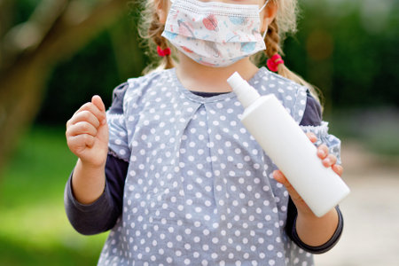 Little toddler girl in medical mask as protection against pandemic coronavirus quarantine disease. Cute child holding bottle with sanitizer for cleaning hands. protective equipment against covidの写真素材