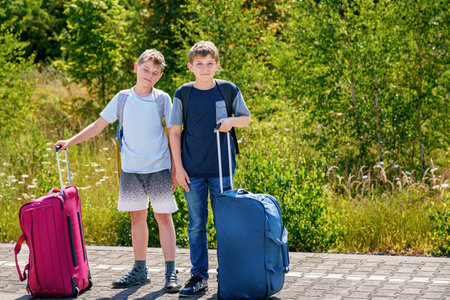 Two children, school boys with suitcases before leaving for summer vacation camp. Happy kids, siblings, twins brothers going on journey, family road trip waiting for train.の写真素材