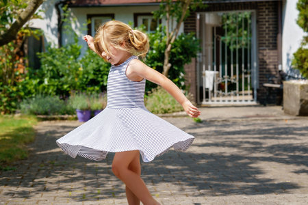Cute little girl in a dress is dancing in garden. Happy child on summer day, outdoors. Carefree childhood.の写真素材