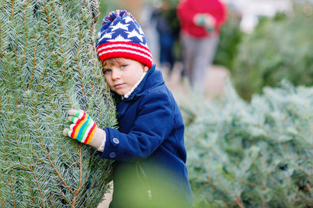 Adorable little smiling kid boy holding Christmas tree on market. Happy healthy child in winter fashion clothes choosing and buying big Xmas tree in outdoor shop. Family, tradition, celebration.の写真素材