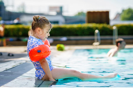 Little preschool girl with protective swimmies playing in outdoor swimming pool by sunset. Child...