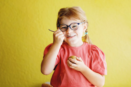 Happy little child girl with glasses eating kiwi fruit with spoon on yellow background. Preschool girl smiling. Healthy fruits for childrenの写真素材