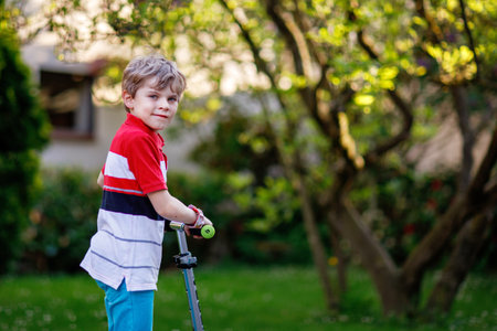 Happy little school kid boy riding on scooter on way to elementary school. Child without safety helmet, school bag on sunny warm morning. Traffic in the city and school children.の写真素材