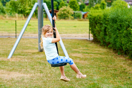 Happy kid girl rids on zip line swing outdoor game play equipment on playground. Child having fun outdoors. Preschool child swinging on summer day. Activity with children.の写真素材