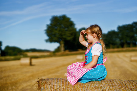 Cute little kid girl in traditional Bavarian costume in wheat field. Happy child with hay bale in Munich. Preschool girl play at hay bales during summer harvest time in Germany.の写真素材