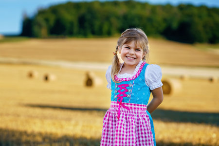 Cute little kid girl in traditional Bavarian costume in wheat field. Happy child with hay bale in Munich. Preschool girl play at hay bales during summer harvest time in Germany.の写真素材