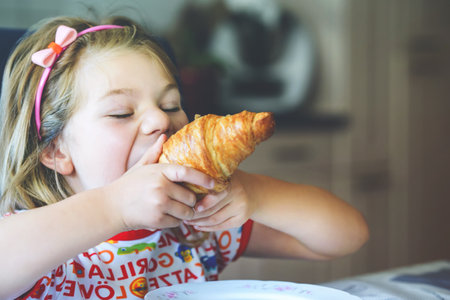Smiling child at breakfast. Food and happy kids. The girl is eating a croissant. Cute preschool girl having healthy meal.の写真素材