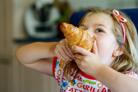 Smiling child at breakfast. Food and happy kids. The girl is eating a croissant. Cute preschool girl having healthy meal.の写真素材
