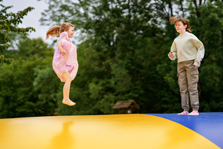Little preschool girl and school sister jumping on trampoline. Happy funny children, siblings in love having fun with outdoor activity in summer. Trampolin in ukrainian flagg colorsの写真素材