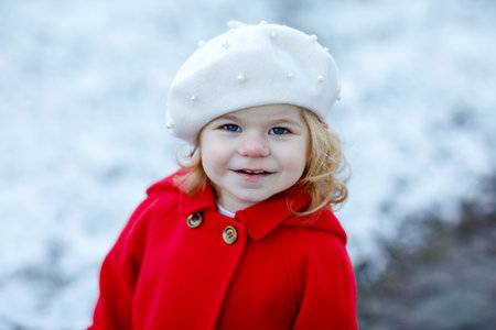Outdoor winter portrait of little cute toddler girl in red coat and white fashion hat barret. Healthy happy baby child walking in the park on cold day with snow and snowfall. Stylish clothes for kids.の写真素材