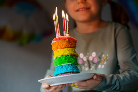 Happy little preschool girl celebrating birthday. Closeup of child with homemade rainbow cake, indoor. Happy healthy toddler blowing six candles on cake. Selective focus on cakeの写真素材