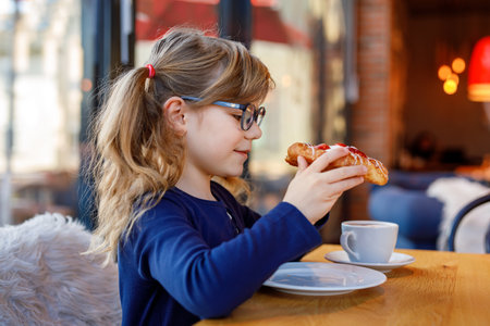 Adorable smiling girl with glasses have a breakfast in a cafe. Preschool child with glasses drinking chocolate and eating bakery pastry croissant or cake. Happy children, healthy food and meal.の写真素材