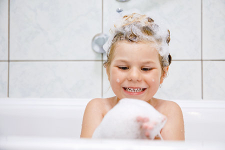 Cute child with shampoo foam and bubbles on hair taking bath. Portrait of happy smiling preschool girl health care and hygiene concept. Washes hair by herself.の写真素材