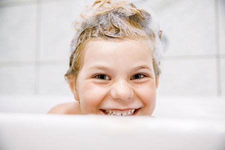 Cute child with shampoo foam and bubbles on hair taking bath. Portrait of happy smiling preschool girl health care and hygiene concept. Washes hair by herself.の写真素材